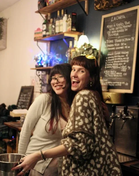 Deux amies éclatent de rire derrière le comptoir, devant l'ardoise et les tireuses à bière